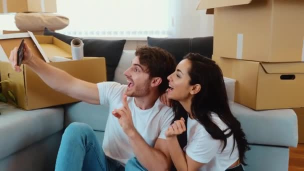 Junges Paar bezieht neue Wohnung Die Kamera bewegt sich rückwärts von Menschen, die auf dem Boden sitzen und Selfies machen. Lustige Mimik und Pose. Glückliche Menschen nach dem Umzug. — Stockvideo