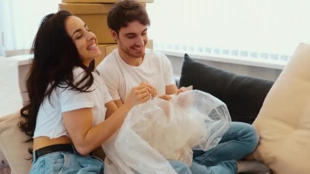 Young couple move into new apartment. Man an woman sit on sofa together and playing with bubble wrap. Rest and relax in middle of relocation into new apartment. — Stock Video