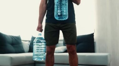Genç adam evde egzersiz yapıyor. Sıkıcı fitness sporcusu birbiri ardına şişe kaldırıyor. Oturma odasında tek başına antrenman yapan ve poz veren adam. Kamera geri gidiyor.
