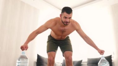 Genç adam evde egzersiz yapıyor. Su şişeleriyle el kaldırma ve kol kaslarıyla çalışma. Güç ve güç kazanmak için antrenman. Su şişelerini bırak ve poz ver..