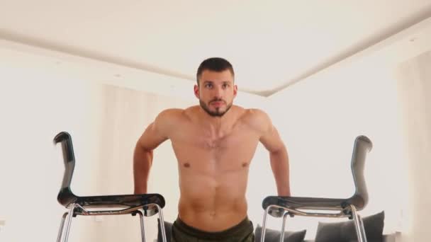 Un joven haciendo ejercicio en casa. Video de un tipo haciendo flexiones verticales usando dos sillas de él. Levantando el cuerpo hacia arriba y hacia abajo. Entrenamiento intensivo potente . — Vídeos de Stock