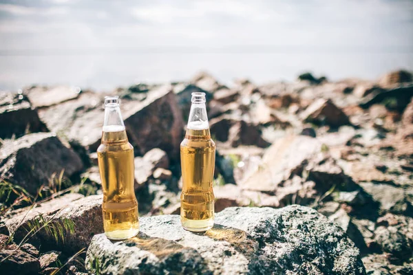 Close-up duas garrafas de cerveja estão em pedras perto da água sob o sol contra o céu — Fotografia de Stock