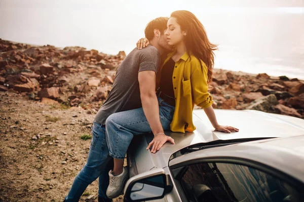 Pareja joven y romántica compartiendo un momento especial al aire libre. Pareja joven enamorada en un viaje por carretera. Pareja abrazándose mientras se sientan en la capucha de su coche en la naturaleza . — Foto de Stock