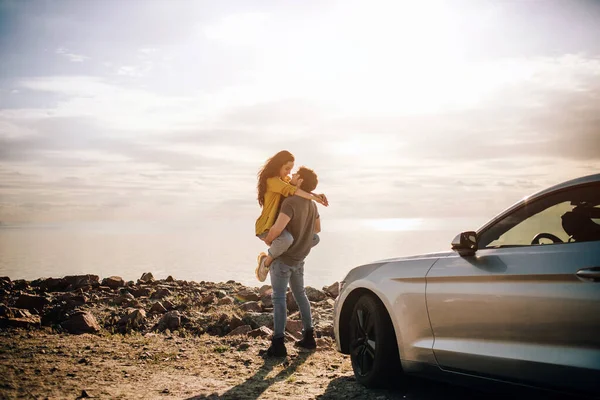 Casal romântico está de pé perto de um carro Muscle na praia. O homem barbudo bonito e uma jovem atraente têm uma história de amor . — Fotografia de Stock