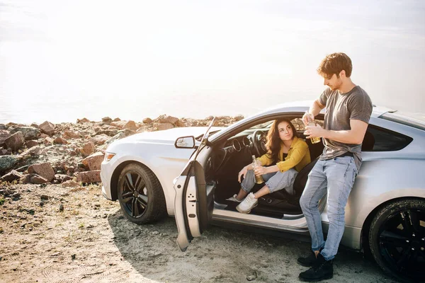 Casal romântico está de pé perto de um carro Muscle na praia. O homem barbudo bonito e uma jovem atraente têm uma história de amor . — Fotografia de Stock