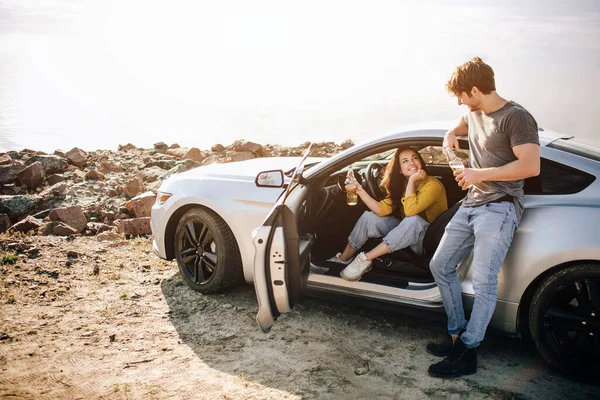 Casal romântico está de pé perto de um carro Muscle na praia. O homem barbudo bonito e uma jovem atraente têm uma história de amor . — Fotografia de Stock