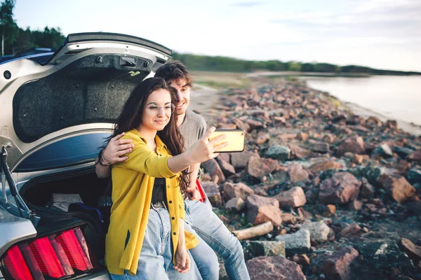Boldog utazó pár ül az autó nyitott csomagtartóban, és nézni a napfelkeltét, és figyelembe Selfie. — Stock Fotó
