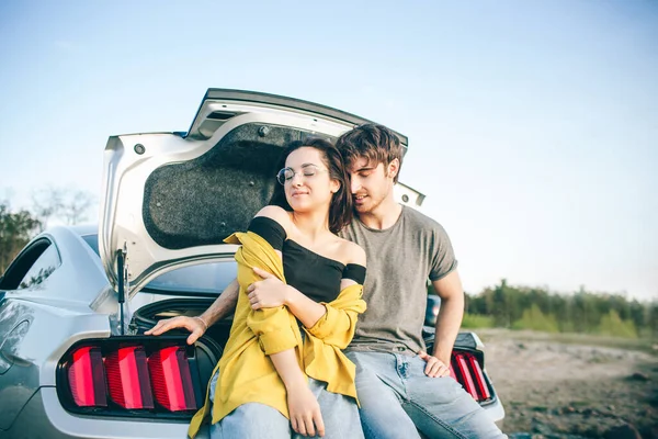 Happy traveler couple sitting in car open trunk and watch the sunrise