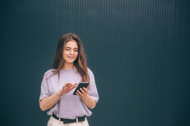 Gri mavi arka planda izole edilmiş modaya uygun genç bir kadın. Kız akıllı telefonu elinde tutuyor ve kullanıyor. İnternette aranıyor. Telefonu kontrol ediyorum. Tek başına dur ve poz ver.