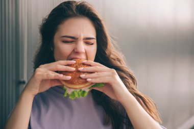 Gri mavi arka planda izole edilmiş modaya uygun genç bir kadın. İki eliyle hamburger tutan ve ısıran bir kız. Öğle yemeğinde nefis yemekler..