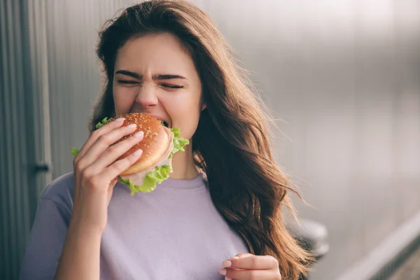 Fiatal, divatos nő elszigetelt szürke kék háttér. Éhes lány, aki hamburgert harap. Ebédidő vagy vacsora. Ízletes hamburger a kézben.. — Stock Fotó