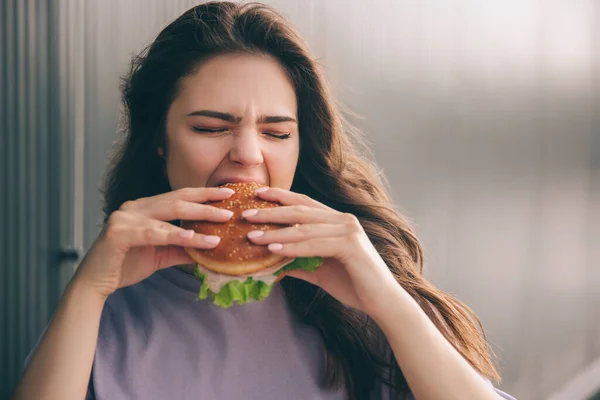 Ung snygg trendig kvinna isolerad över grå blå bakgrund. Flicka som håller i hamburgare med två händer och biter en bit av den. Smaklig läcker måltid under lunchtid. — Stockfoto