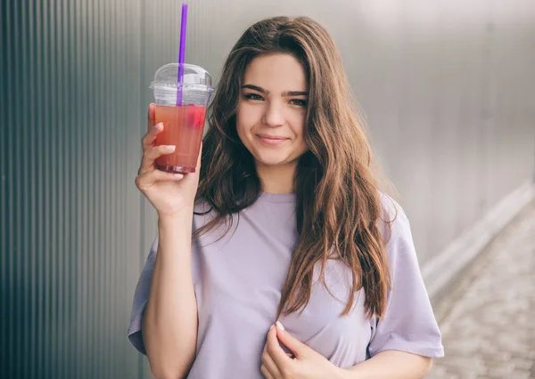 Young stylish trendy woman isolated over grey blue background. Cheerful positive girl posing on camera with plastic cup in hand. Drinking red lemonade. Lovely girl looks straight. Alone outside.