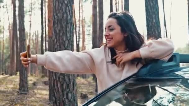 Молода жінка їде за межами машини в лісі і записує відео за допомогою смартфона. Блогер створює відеоконтент. Рухається рука з телефоном в ньому і позує з посмішкою . — стокове відео