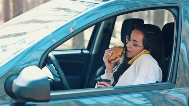 Jonge vrouw zit in de auto tijdens de reis pauze. Meisje eet hamburger en drinkt uit thermoskan. Telefonisch praten en een gesprek voeren. Online communicatie. Langzame beweging. — Stockvideo