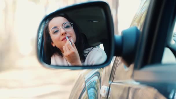 Mujer joven sentada en coche durante la pausa de viaje. Atractiva chica hermosa mirando en el espejo lateral y la aplicación de lápiz labial rosa en los labios. Cuida de su maquillaje. Tiempo de cosméticos . — Vídeos de Stock