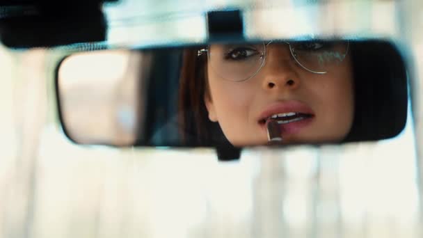 Молодая женщина сидит в машине во время перерыва. Девушка наносит красную или обнаженную помаду на губы и смотрит в зеркало обзора. Улыбаясь своему отражению и глядя в камеру . — стоковое видео