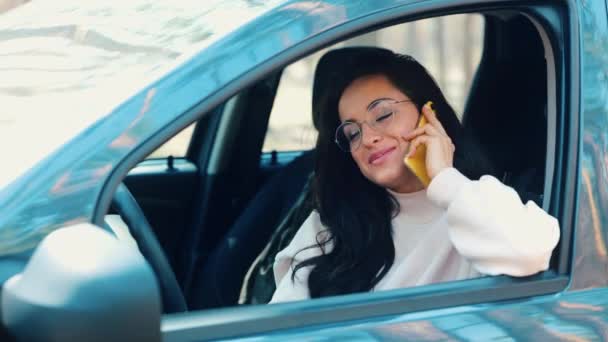 Молодая женщина сидит в машине во время перерыва. Восхитительная позитивная девушка разговаривает по телефону и улыбается. Сидя внутри во время перерыва и наслаждаясь телефонным разговором . — стоковое видео
