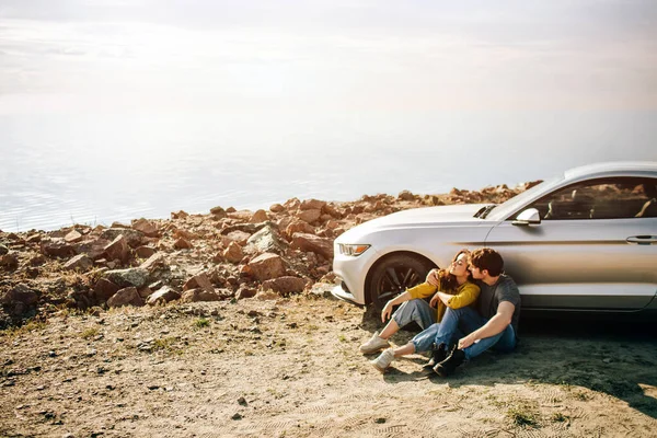 Casal romântico está de pé perto de um carro Muscle na praia. O homem barbudo bonito e uma jovem atraente têm uma história de amor . — Fotografia de Stock