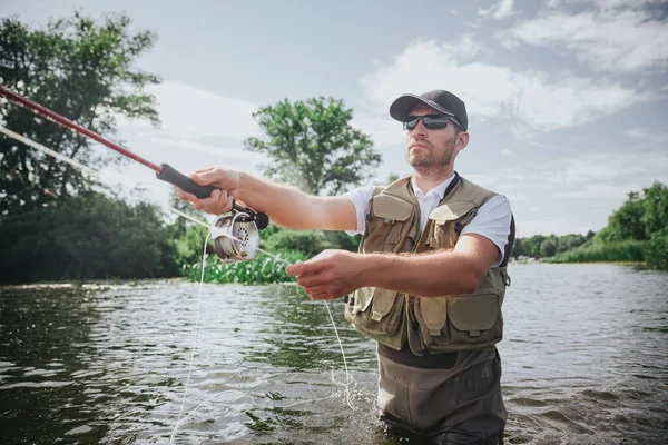 Ung fiskare som fiskar på sjö eller flod. Koncentrerad kille som håller i spö med fiskelina i händerna och kastar den för att fånga fisk. Stå i mitten av vatten flod eller sjö. — Stockfoto