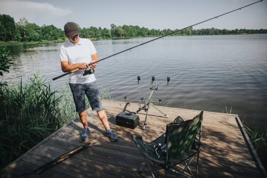 Gölde ya da nehirde balık tutan genç bir balıkçı. Olta makarasını kullanarak ve onu ayarlayarak balık tutmaya hazırlanıyorum. Nehir kıyısında uzun bir sopayla tek başına dur..