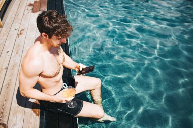 Genç seksi adam yüzme havuzunda dinleniyor. Tepede oturan ve suyun içinde bacakları tutan adamın manzarası. Akıllı telefon kullanan adam. Tatil için spa merkezinde zayıf, yapılı, güçlü bir adam..