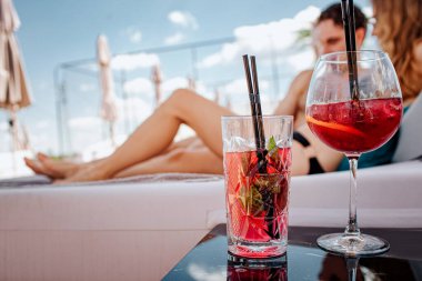 Genç ve seksi bir çift yüzme havuzunda dinleniyor. Masada iki kırmızı, soğuk, lezzetli kokteyl duruyor. Erkek ve kadın yaz tatili boyunca tatil köyünde uzanıp dinleniyorlar. Hava ve arkadaşlığın tadını çıkar.