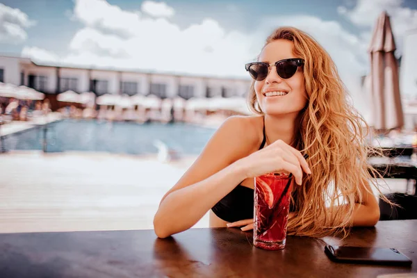 Young hot woman resting at swimpool. Gorgeous girl sitting alone at bar and enjoying drinking cocktail. Sexy woman in black swimsuit rest in resort place.