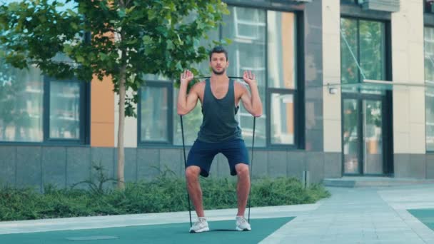 운동하는 젊은이. 활기차고 강한 근육을 가진 남자의 느린 움직임을 보고 있습니다. 고무줄을 사용하여 웅크리기 운동을 합니다. 거리에서 운동을 하는 훈련. — 비디오