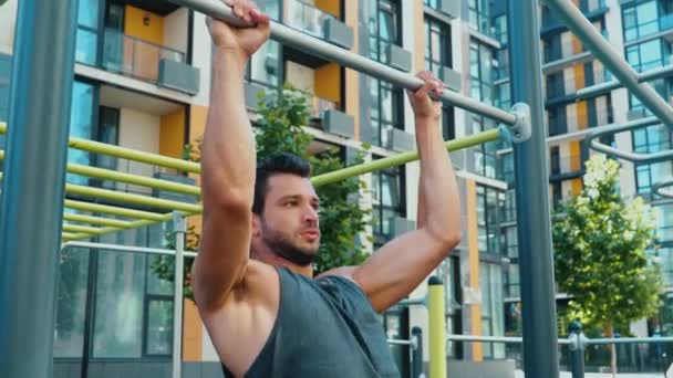 Jongeman die buiten traint. Langzame beweging en achteraanzicht van gespierde sterke krachtige man doen pull-up oefening door opknoping op horizontale balk. Training op straat in een stedelijk gebouw. — Stockvideo