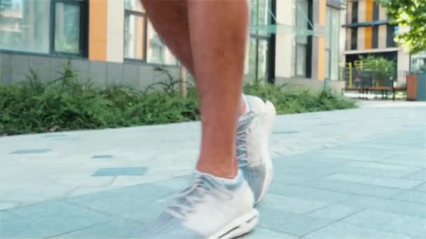 Junger Mann beim Training draußen. Seitenansicht Zeitlupe von Jungs dünne Füße in weißen Turnschuhen joggen oder langsam auf der Straße am Gebäude laufen. Bewegungstraining für Gesundheit und Vitalität. — Stockvideo
