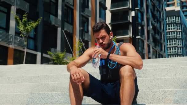 Jeune homme faisant de l'exercice dehors. Fatigué gars épuisé assis sur les marches sur la rue et boire de l'eau. Une corde sautante pend autour de son cou. Après avoir couru ou fait du jogging dehors. — Video