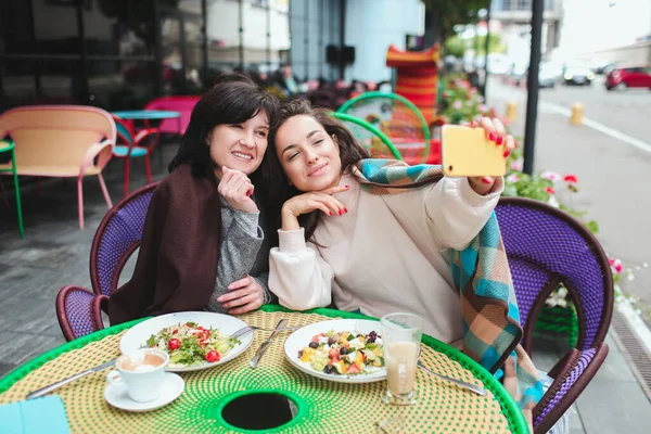 Olgun anne ve küçük kızı kafede ya da restoranda birlikte oturuyorlar. Kız annesiyle selfie çekiyor. Kafede oturup kameraya poz vermek. Dışarıda iki kadının resmi var. İyi bir ilişki. — Stok fotoğraf