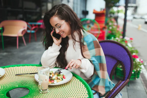 Молода жінка сидить сама в кафе або ресторані. Красива позитивна дівчина розмовляє по телефону під час обіду. Сидячи за столом і їдячи салат. Один назовні . — стокове фото