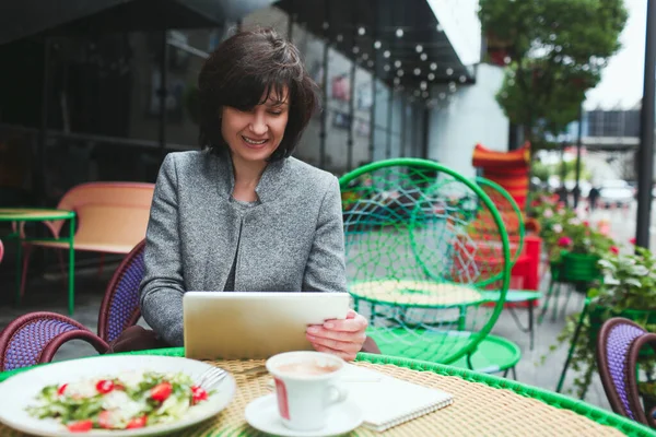 Mature woman sit alone in cafe or restaurant. Adult businesswoman hold tablet and work on it. Sit at table during lunch tinme eating salad. Working on project alone.