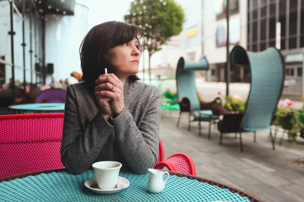 Calm peaceful mature female business woman sitting alone at cafes table and drink coffee. Hold electronic cigarette in hand and look to side. Business lunchtime alone.