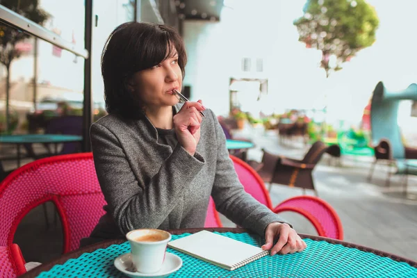 Femme d'affaires mature assis seul et pensant. Prendre des notes dans un carnet. Toucher les dents avec le stylo et regarder de côté. Création d'un nouveau plan d'affaires ou projet. — Photo