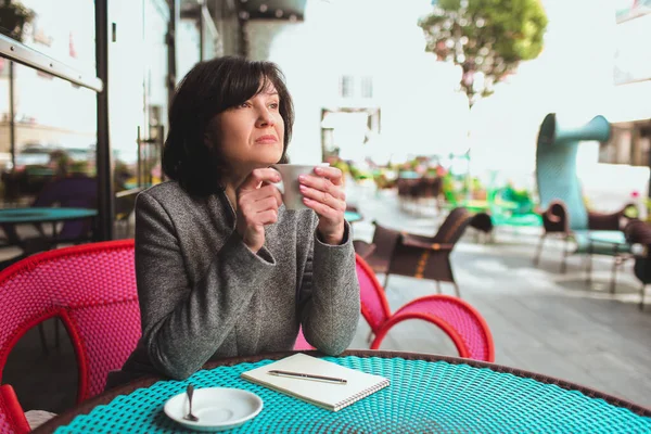 Femme d'affaires mature assis seul et pensant. Tenant une tasse blanche de thé ou de café et regarder de côté. Penser pendant une courte pause. Stylo avec bloc-notes couché sur la table. Lumière du jour dehors. — Photo