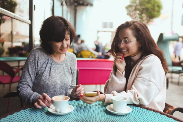 Зрелая мать и ее маленькая дочь сидят вместе в кафе или ресторане. Девушка держит смартфон и показывает что-то на экране своей маме. Зрелая женщина смотрит его. Питьевой чай или кофе. — стоковое фото
