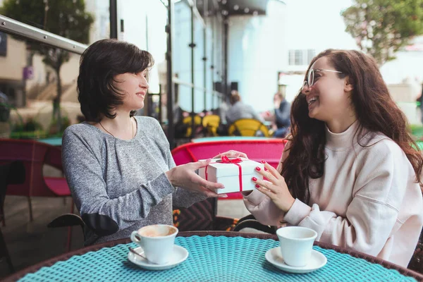 Mère mature et sa jeune fille s'assoient ensemble dans un café ou un restaurant. Une fille donne un cadeau à sa mère. Boîte blanche à rayures rouges. Cadeau ou surprise pour maman mature. Profitez de boire du café dehors. — Photo