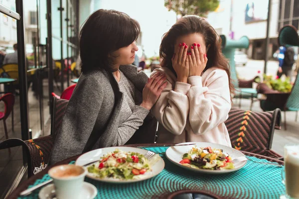 Reife Mutter und ihre kleine Tochter sitzen zusammen im Café oder Restaurant. Mädchen bedecken Gesicht mit Händen und Krallen. Ihre Mutter macht sich Sorgen. Probleme oder Stress. Der Versuch, sich zu beruhigen und besser zu werden. — Stockfoto