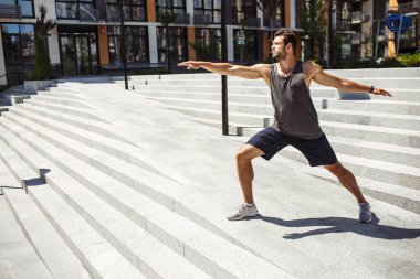 Genç adam dışarıda egzersiz yapıyor. Yoga pozisyonunda tek başına duran ve ellerini ileri geri uzatan adam. Cesetle ilgilenin. Şehir eğitimi. Harika bir refah.