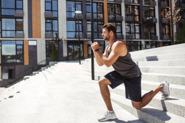 Genç adam dışarıda egzersiz yapıyor. Sporcu sporcunun bir bacağının üzerine çömelmiş bir şekilde aşağıya inişi. Şehir binasının dışında eğitim. Ellerinizi birleştirin ve egzersize konsantre olun..