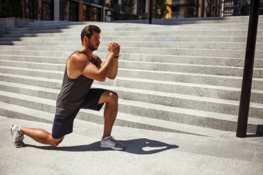 Genç adam dışarıda egzersiz yapıyor. Güçlü ve güçlü bir adam tek dizinin üzerinde duruyor ve önünde el ele tutuşuyor. Bir bacak egzersizi yapıyorum. Vücudunu ve kaslarını çalıştırıyor..