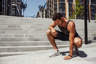 Genç adam dışarıda egzersiz yapıyor. Ciddi bir adam asfalta dokunuyor ve aşağıya bakıyor. Egzersiz, koşu ya da koşudan sonra dinlenin. Basamakların yanında tek başına otur..