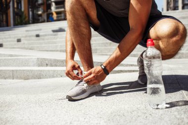 Genç adam dışarıda egzersiz yapıyor. Erkek bağcıklarını, ayakkabı bağcıklarını ya da spor ayakkabılarını kes. Asfalt dışında neredeyse boş bir su şişesi. Sporcu sporcu egzersiz, antrenman ya da spor yapmaya hazırlanıyor..