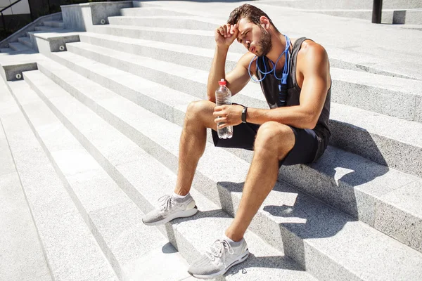 Un joven haciendo ejercicio afuera. Atleta sentarse en las escaleras y descansar después del entrenamiento. Sostiene una botella de agua. Tener una cuerda saltando alrededor del cuello. Tiempo de relajación después del ejercicio. — Foto de Stock