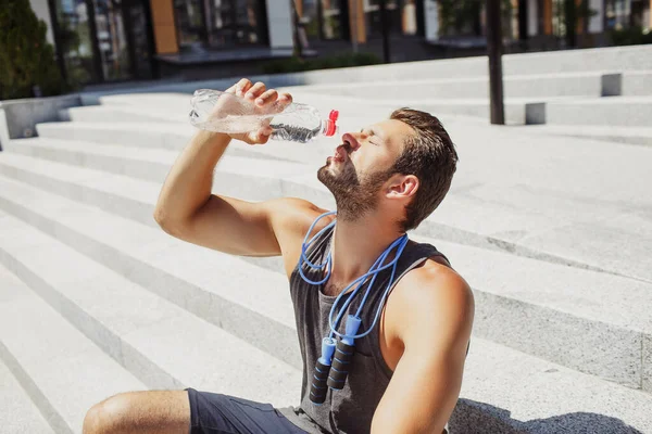 外で運動してる若い男。陸上競技者は一人で階段や飲料水に座っています。彼の体と顔につけて。ハードワークアウト後に休息とリラックス。外のサンシャイン. — ストック写真
