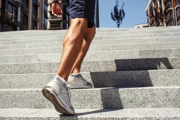Young man exercising outside. Cut view of strong powerful legs and feet in sneakers. Muscled calf. Man walking up on steps at urban building. One more step. Summertime outside.