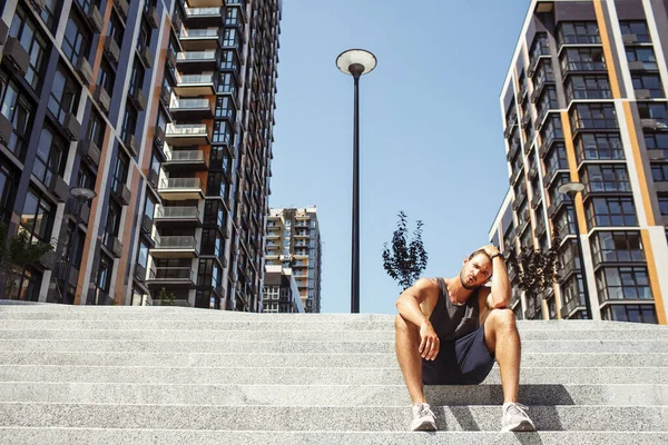 Ung man som tränar utanför. Trött utmattad idrottsman sitter ensam på trappor utanför stadsvyn och vilar efter runing eller hård träning. Sköt om dig. Sitt på huk position. — Stockfoto